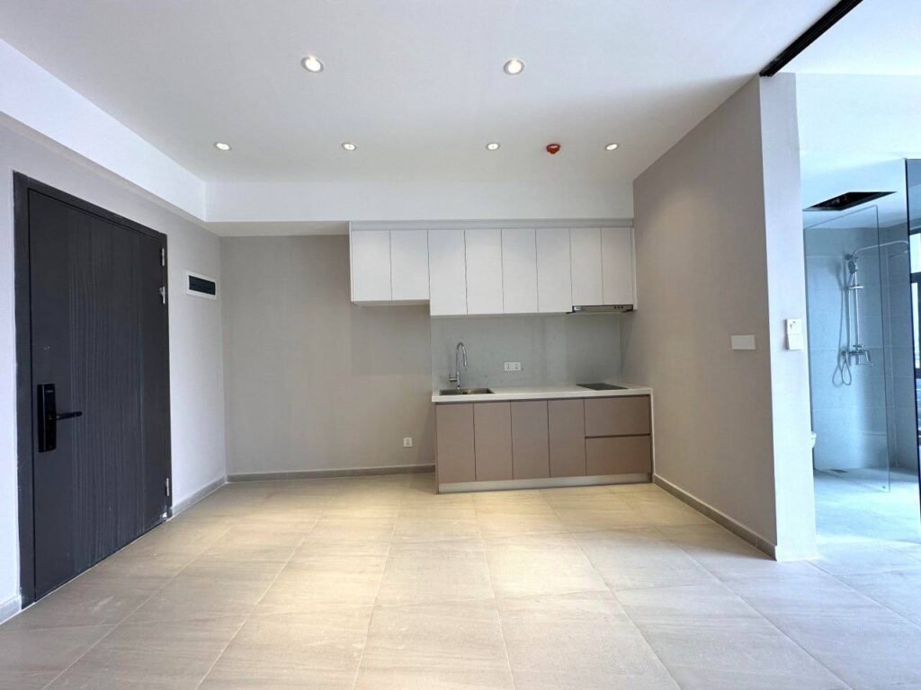 A modern, unfurnished studio apartment in Time Square 3 featuring light-colored tiled flooring, a small kitchenette with white cabinets, a dark wooden door, and a glass-walled bathroom on the right. Ceiling spotlights brighten up the space.