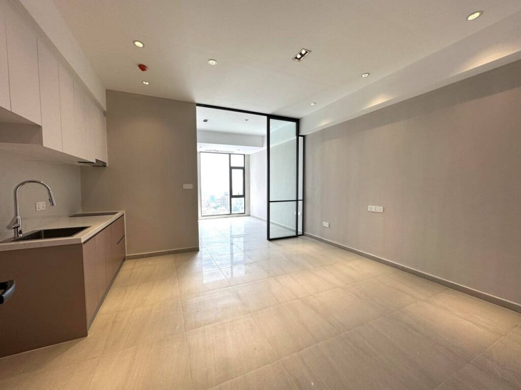 A modern, unfurnished apartment in Time Square 3, featuring beige tile flooring, light gray walls, a compact kitchen with sink and cabinets, large windows, and a glass door opening to a bright room with city views.