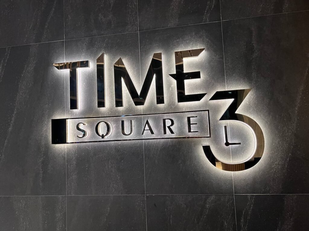 A modern, illuminated wall sign displays TIME SQUARE 3 in bold, metallic letters against a dark, textured background. The prominent number 3 partially overlaps the word SQUARE, highlighting the unique branding of time square 3.