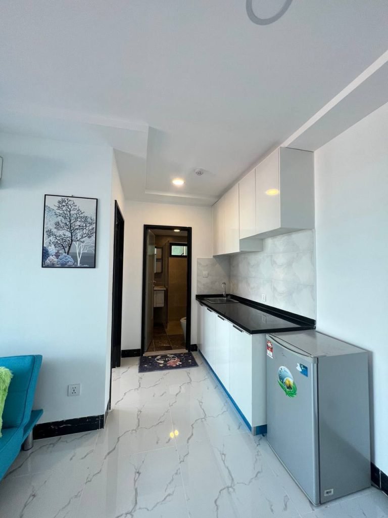 A small modern kitchen with white cabinets, a black countertop, a gray mini fridge, and marble-patterned floor. A doorway leads to a bathroom, and theres wall art of a tree above a blue sofa.