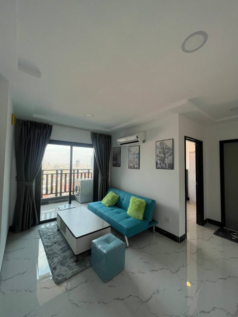 Modern living room with a blue sofa, green pillows, white coffee table, small blue ottoman, and large window with black curtains overlooking a city view. White tile floors and minimal wall art complete the space.