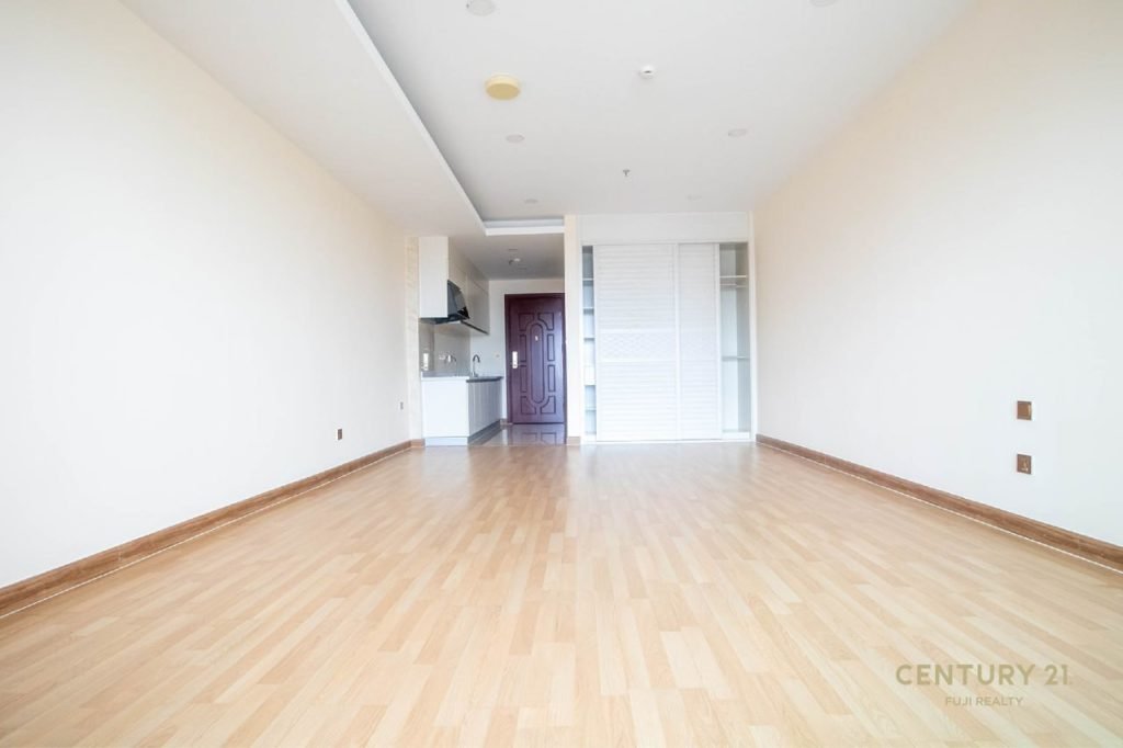 A spacious, unfurnished room with light wood flooring, off-white walls, a visible kitchen area, a brown entrance door, and built-in white storage cabinets.