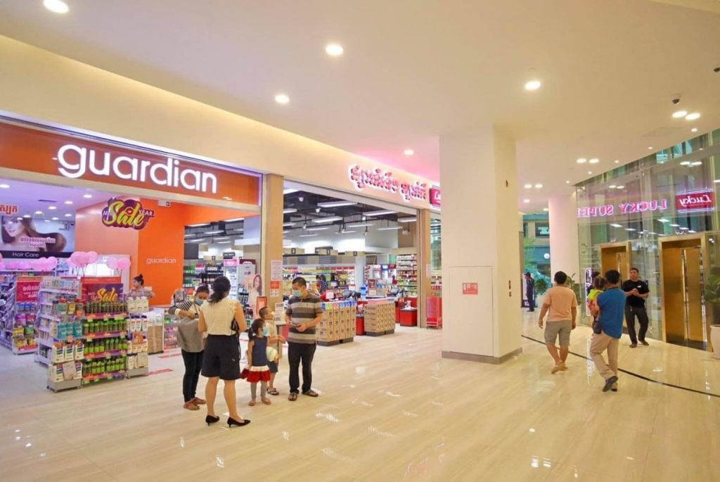 People walk inside a brightly lit shopping mall corridor with a Guardian pharmacy on the left and glass doors on the right. Several shoppers are browsing products near store entrances.