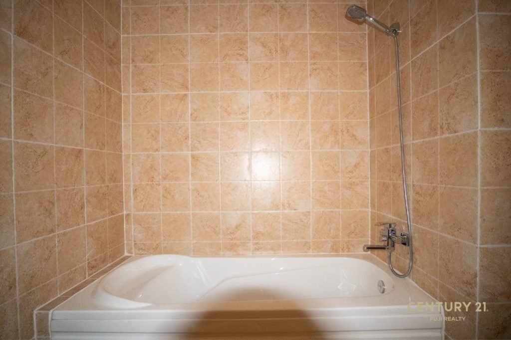 A white bathtub with a wall-mounted showerhead is set against tan, square tiled walls in a bathroom. The tiles have a warm, earthy tone, and the shower fixtures are chrome.