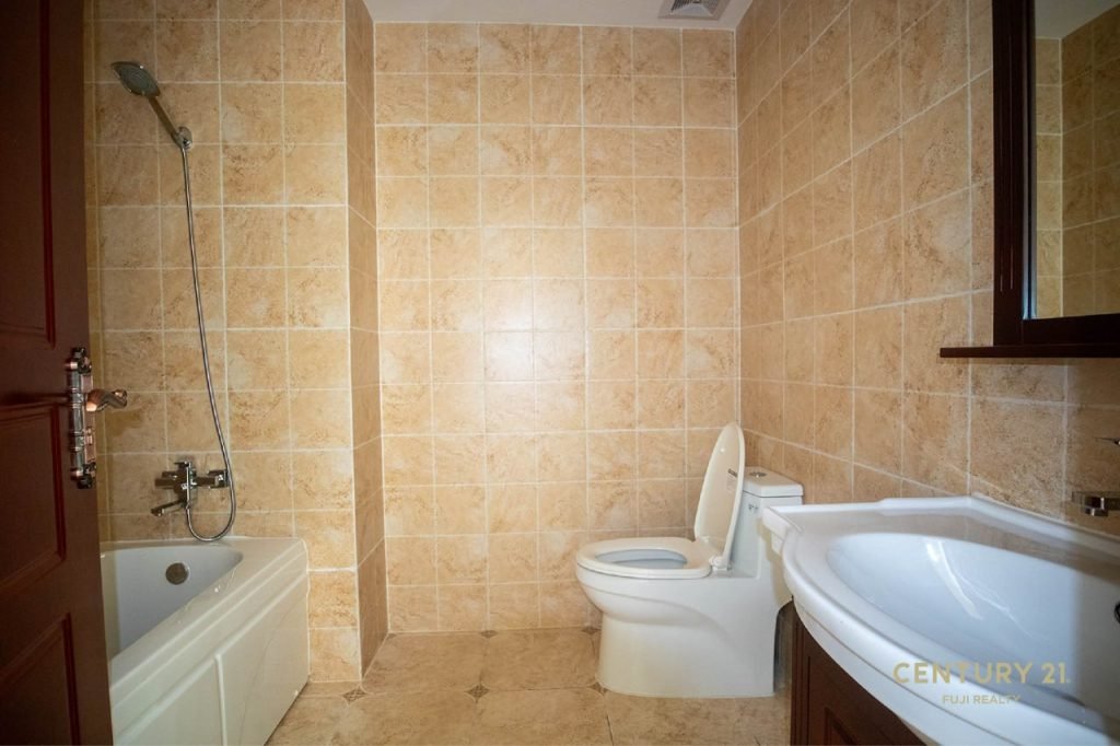 A bathroom with beige tiled walls and floor, featuring a white bathtub with a showerhead, a toilet, and a white sink with a mirrored cabinet.