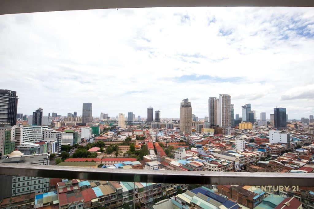 A cityscape view from a high-rise balcony at Olympia city, Phnom Penh, showing densely packed buildings, skyscrapers, and cloudy sky above.