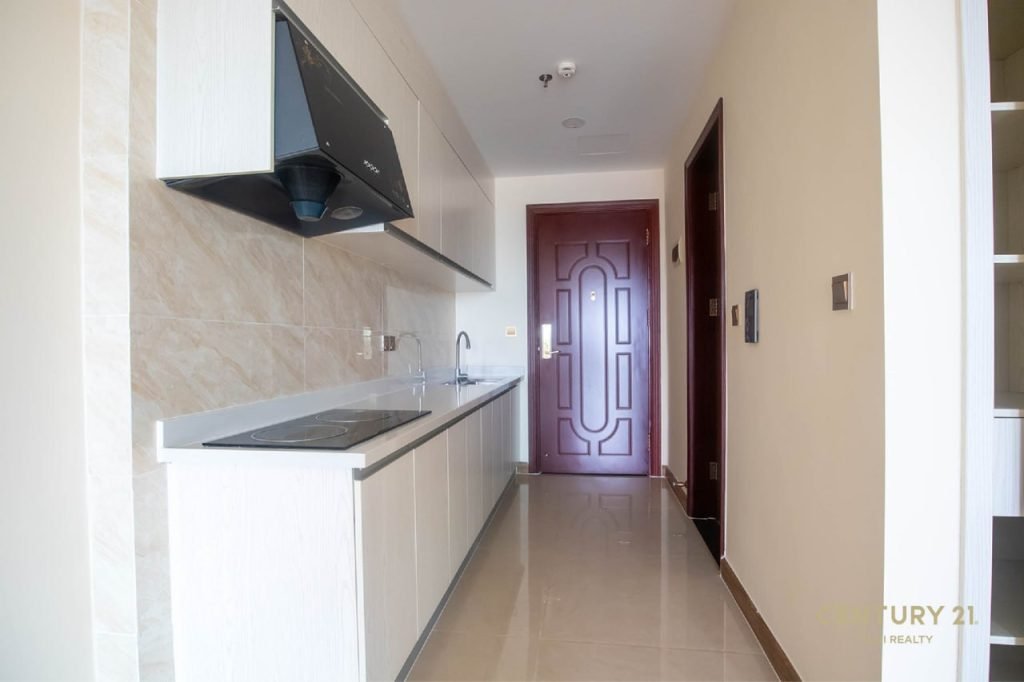 A narrow, modern kitchen with white cabinets, a sink, a stovetop, beige tiled walls, and glossy floor tiles. A red door is at the end of the hallway.