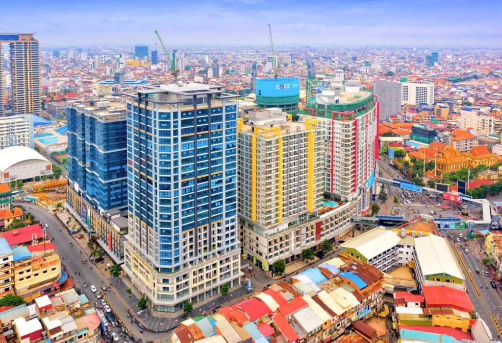 Aerial view of Phnom Penh with tall modern buildings, colorful rooftops, busy roads, and dense urban development stretching into the distance under a partly cloudy sky.