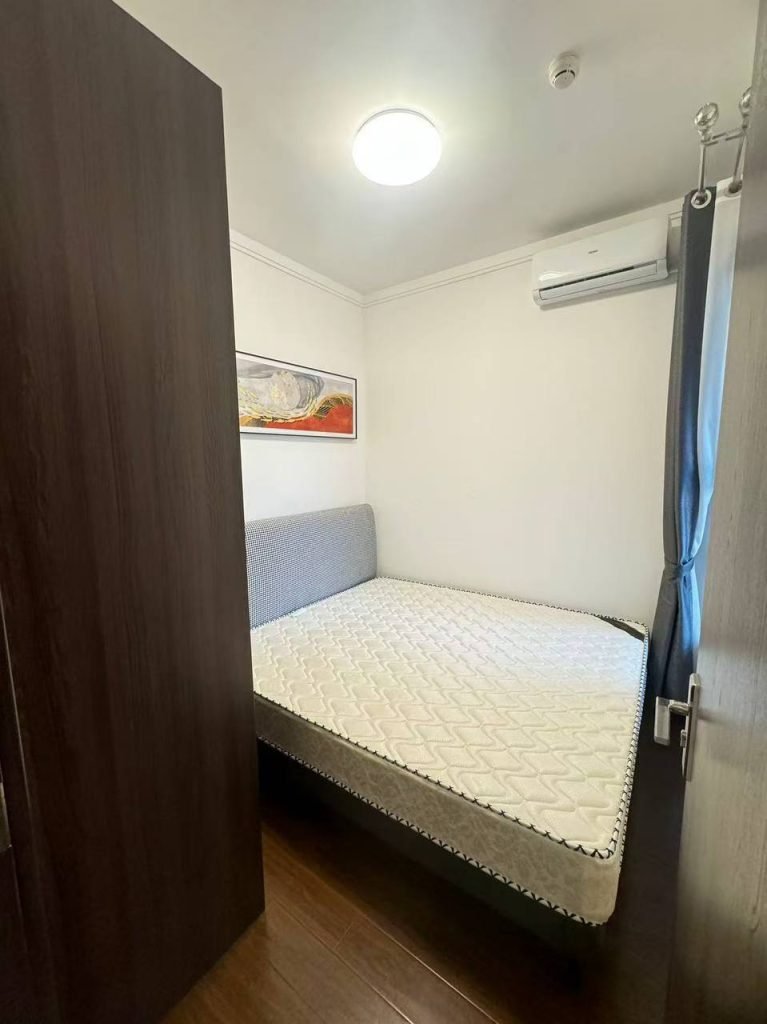 A small, tidy bedroom with a double bed, grey upholstered headboard, white mattress, wall-mounted air conditioner, abstract artwork on the wall, dark wooden floor, and a window with dark curtains.