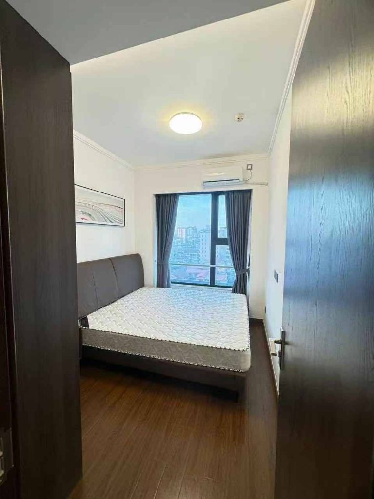 A small, modern bedroom with a double bed, dark headboard, wooden floor, large window with city view, blue curtains, air conditioner, and a round ceiling light. The door is partially open.