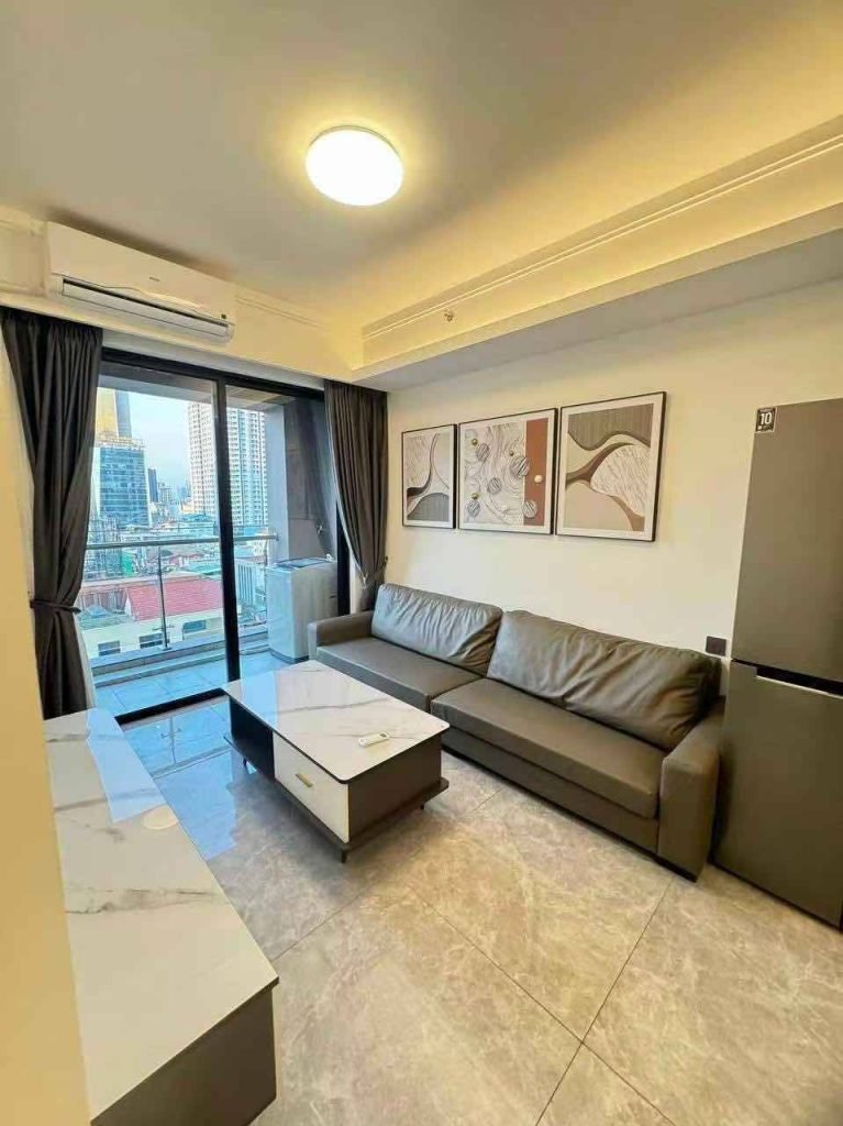 Modern living room with a gray sofa, marble coffee table, wall art, and a refrigerator. Large window with city view, balcony, and dark curtains. Light-colored tiled floor and air conditioner are visible.