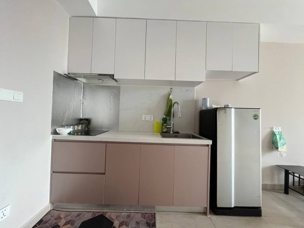 A compact modern kitchen with upper white cabinets, lower brown cabinets, a stainless steel sink, a small refrigerator, a silver backsplash, and a bowl on the counter. A green towel hangs above the sink.