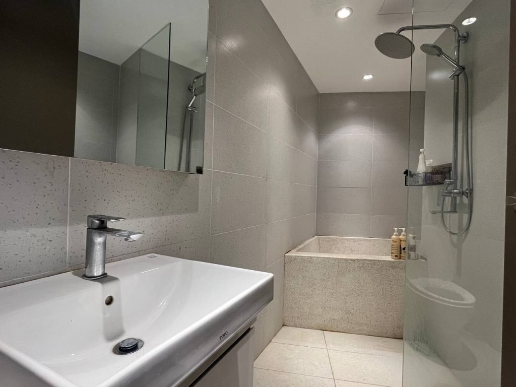 Modern bathroom with a white rectangular sink, wall-mounted faucet, large mirror, grey tiled walls, and a walk-in shower next to a deep soaking tub with toiletries on the ledge. Bright ceiling lights illuminate the space.