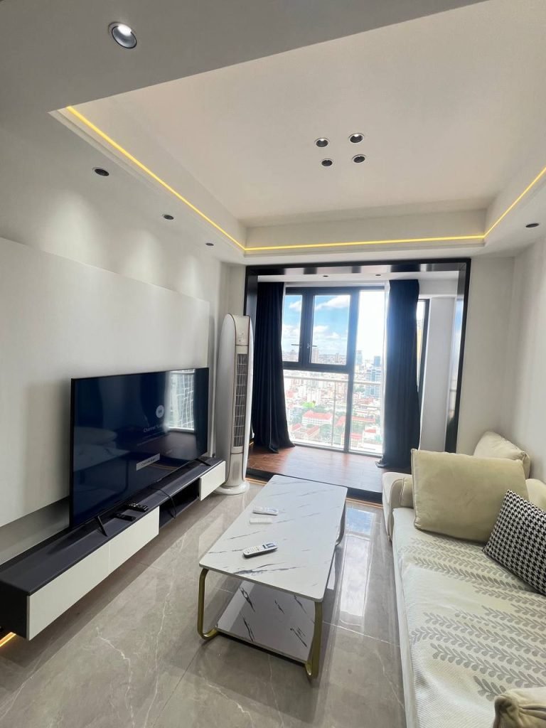 Modern living room with a large wall-mounted TV, white and beige sofa, marble coffee table, TV stand, and floor-to-ceiling window with city view; neutral tones and recessed ceiling lighting.