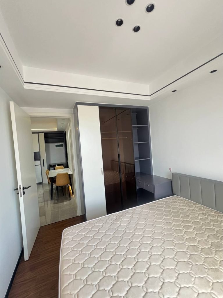 A modern, bright bedroom with a bed, built-in wardrobe with glass doors, shelves, and a small desk. An open door reveals a dining area with a table and chairs in the background.