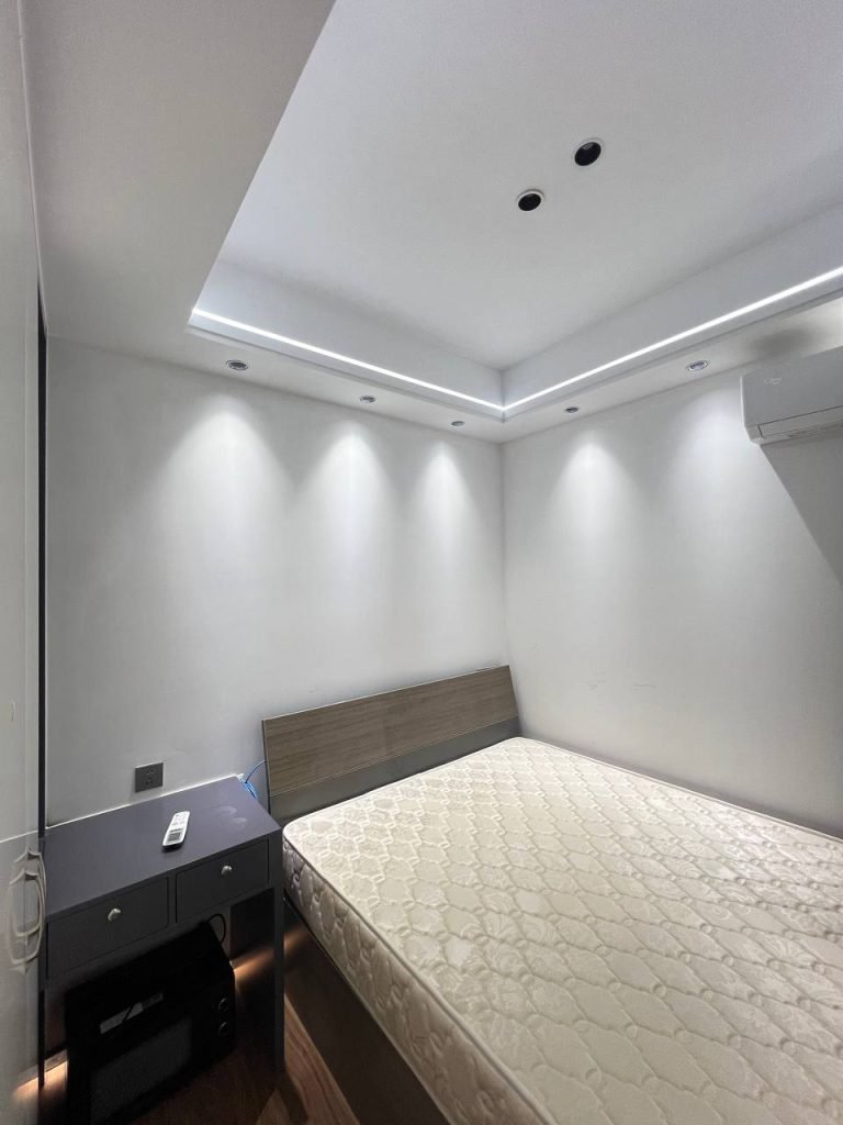Minimalist bedroom with a large bed, plain white walls, recessed ceiling lights, a small dark-colored desk with a drawer, and an air conditioner mounted on the wall. The room appears neat and uncluttered.