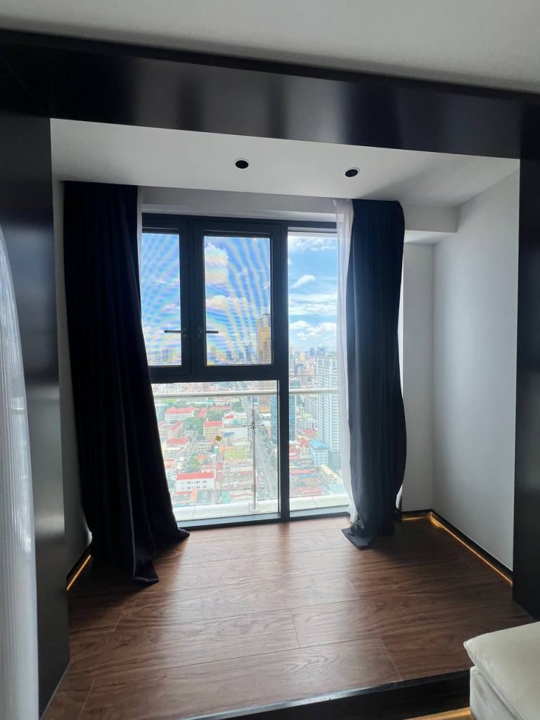 A modern room with wooden flooring, black curtains partly open, and a large window overlooking a cityscape under a blue sky with clouds. Natural light fills the space, highlighting the urban view outside.