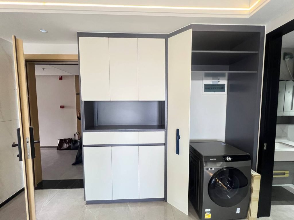Modern laundry area with a black washing machine beneath open shelving, adjacent to built-in cabinets with a recessed shelf; a door opens to a hallway with a bag on the floor in the background.