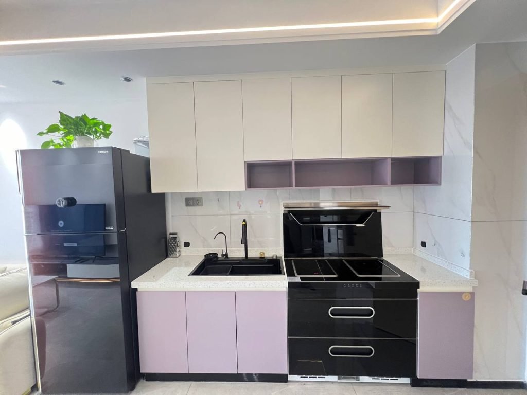 A modern kitchen with purple and white cabinets, a black refrigerator, a black electric stove and oven, a black sink, and a potted plant on top of the fridge. Marble backsplash and minimalist design.