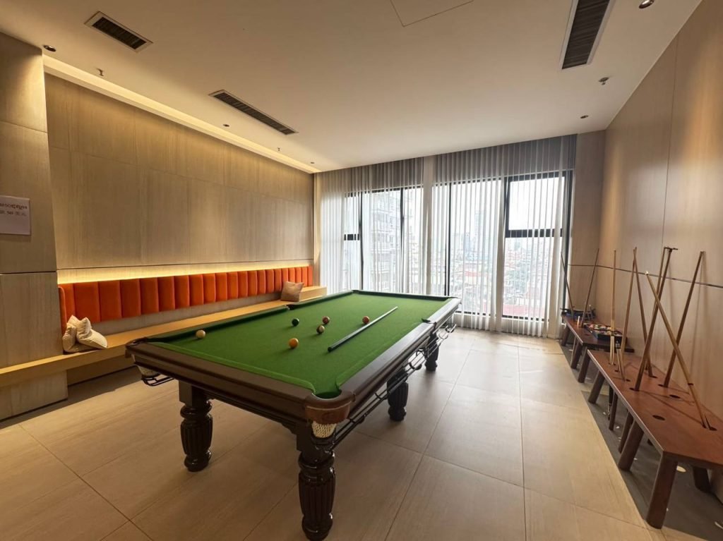 A modern room with a green snooker table in the center, balls arranged on the table, cues resting on a rack, a long orange-cushioned bench along one wall, and large windows with sheer curtains letting in daylight.