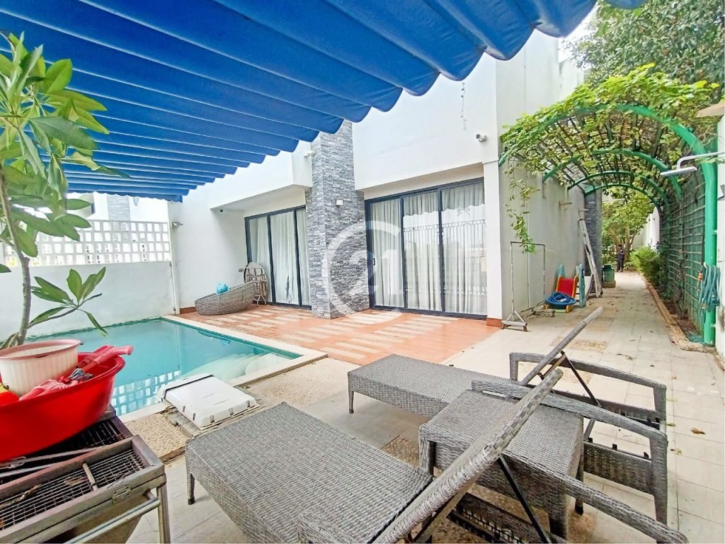 A modern backyard features a small swimming pool, lounge chairs, a blue awning, potted plants, and a vine-covered green archway leading to a side path beside a white building with large glass doors.