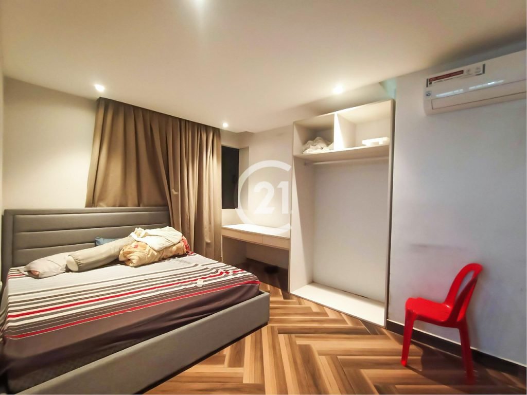 A small bedroom with a striped bed, brown curtains, a built-in closet with shelves, a red plastic chair, a white air conditioner, and wood-patterned flooring under warm ceiling lights.