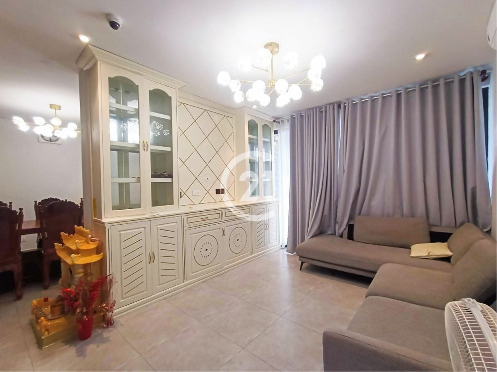 A modern living room with light gray floor tiles, gray sofas, a white built-in cabinet, and gray curtains. A chandelier hangs from the ceiling. There is a small decorative altar with red and gold elements in the corner.