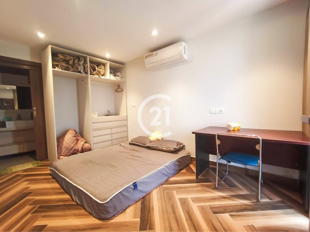 A minimalist bedroom with a mattress on the wooden floor, a black desk with a chair, built-in shelves with folded bedding, and an air conditioner above. The room is well-lit, with light-colored walls and modern decor.