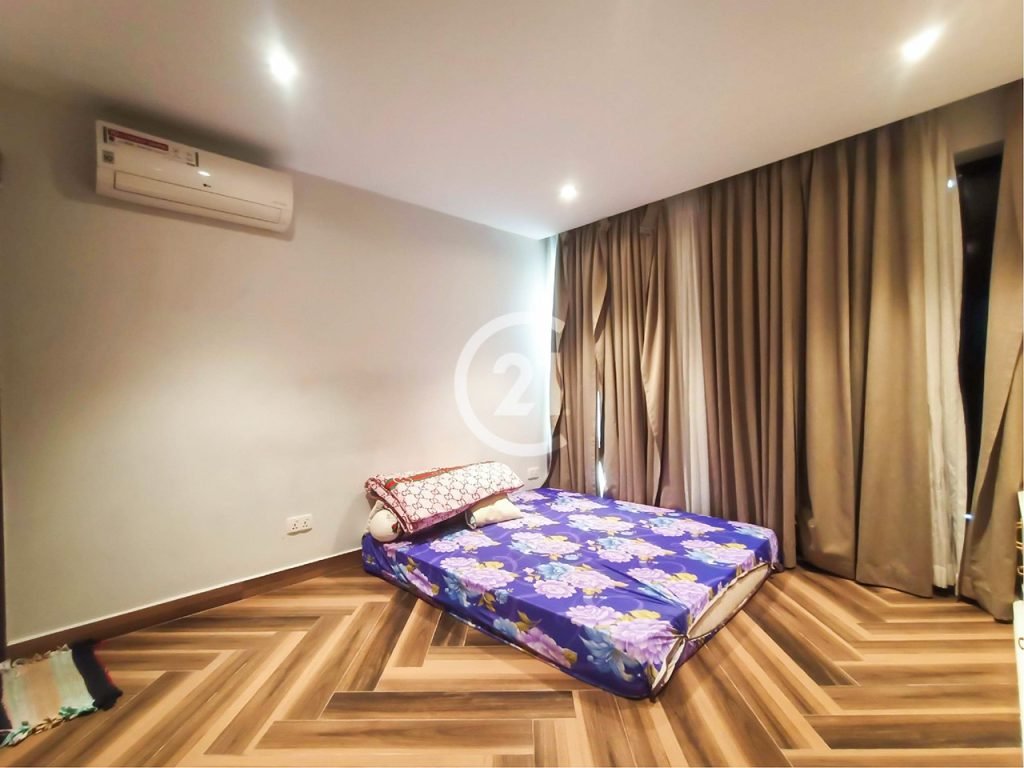 A minimalist bedroom with a floral-patterned mattress on the wooden herringbone floor, beige curtains covering large windows, white walls, an air conditioner, and soft ceiling lights.