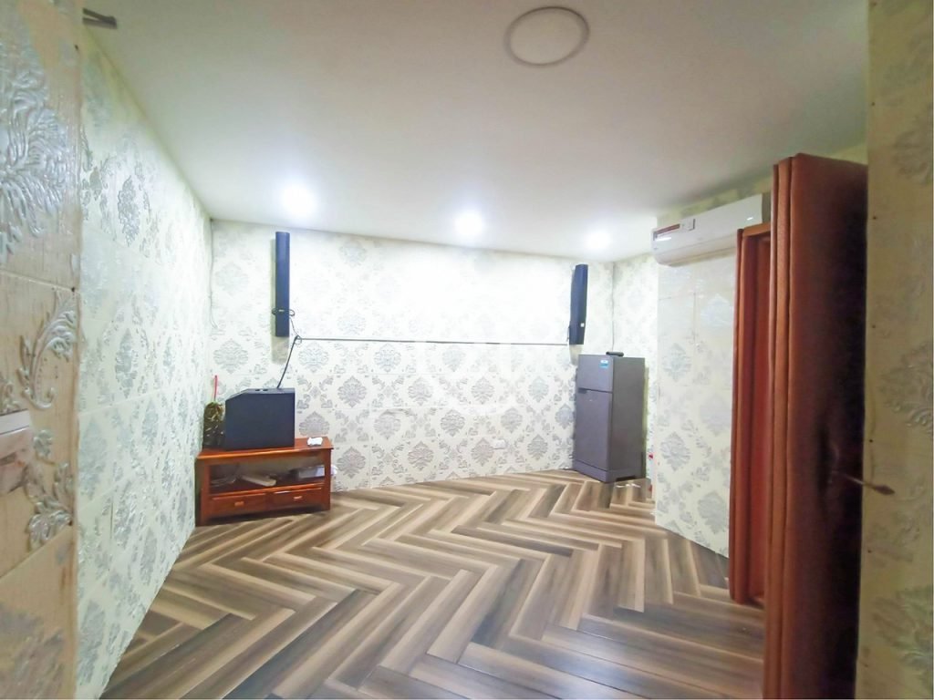 A room with patterned wallpaper, wooden herringbone flooring, a TV on a wooden stand, speakers mounted on the wall, a refrigerator, and an air conditioner above a door. Ceiling lights are on.