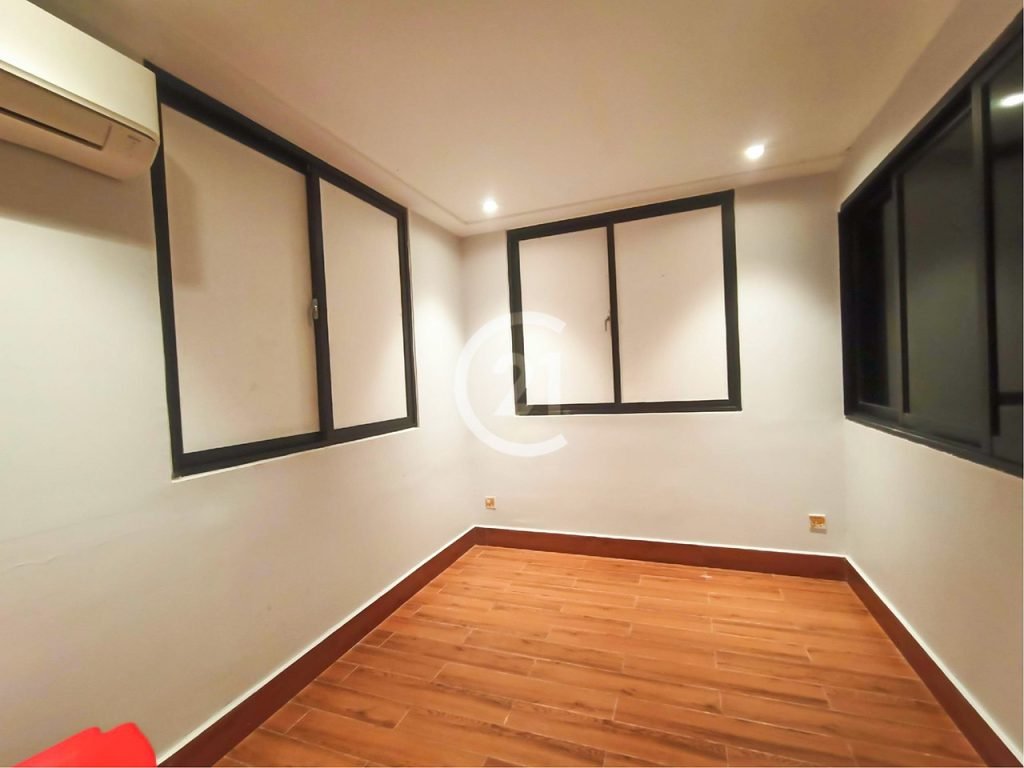 A small, empty room with white walls, brown wooden floors, black-framed windows, an air conditioner on the left wall, and recessed ceiling lights.