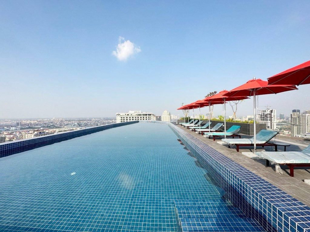 Rooftop infinity pool with blue tiles, lined with lounge chairs and red umbrellas, overlooking a cityscape under a clear, sunny sky.