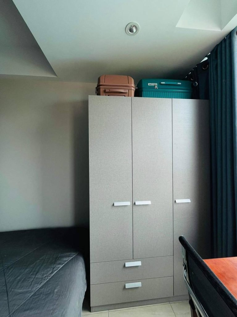 A gray wardrobe with three doors stands in a corner next to a bed with a gray blanket. Two suitcases, one pink and one teal, are on top of the wardrobe. A desk and curtain are partially visible on the right.