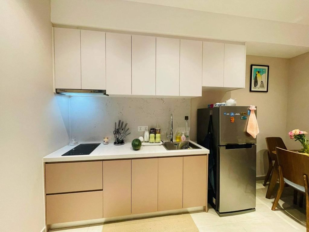 A modern kitchen with pale pink lower cabinets, white upper cabinets, an induction cooktop, utensils, a sink, and a stainless steel refrigerator. A small dining table with flowers is visible on the right.