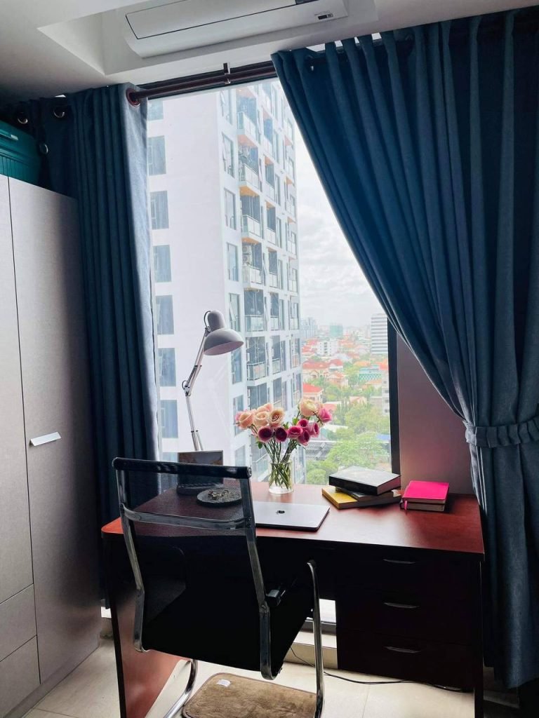 A modern home office with a dark desk facing a large window, blue curtains, a black chair, a laptop, books, a desk lamp, and a vase of flowers, overlooking a cityscape with high-rise buildings.