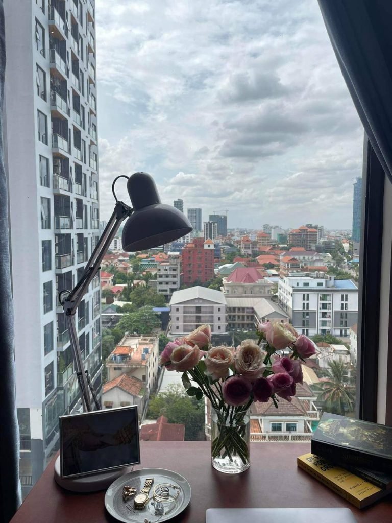 A modern desk with a lamp, a vase of pink flowers, a framed photo, jewelry, and books sits by a window overlooking a cityscape with tall buildings and a cloudy sky.