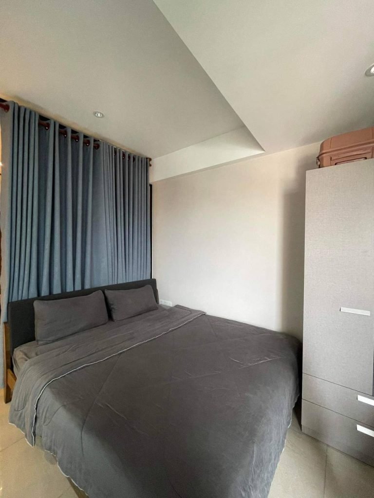 A modern bedroom with a large bed covered in grey bedding, grey curtains along the wall, a light-colored wardrobe with drawers on the right, and a pink suitcase on top of the wardrobe. The room is minimal and tidy.