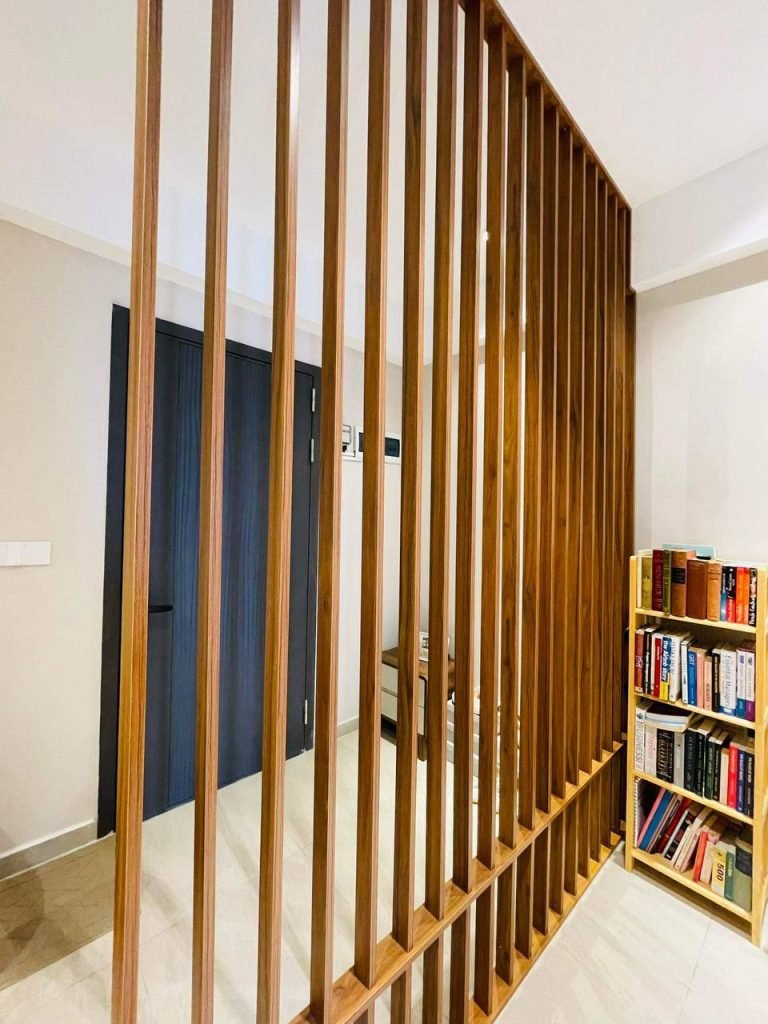 A modern interior featuring a vertical wooden slat room divider, a bookshelf filled with books on the right, and dark-colored doors in the background. The space is bright with neutral-colored walls and flooring.
