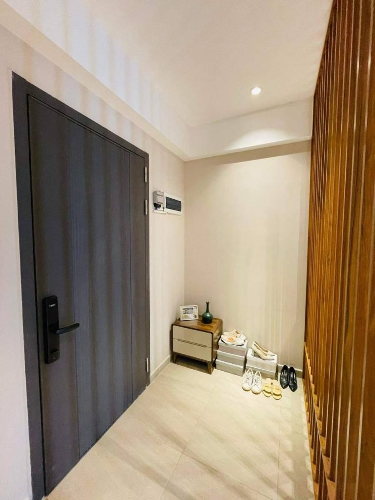A modern entryway with a dark door, light walls, a small wooden table holding decor items, several pairs of shoes on the floor, and wooden slats on the right side. The area is well-lit with a ceiling light.