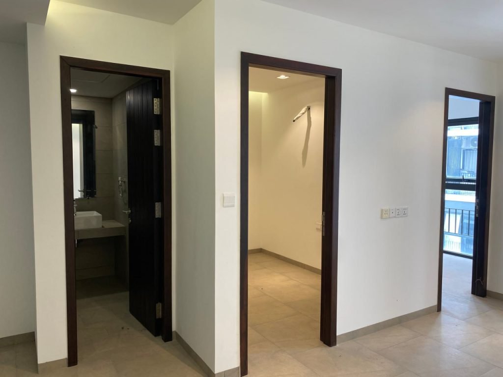 A bright, empty room with beige tiled flooring. Two open doorways are visible: one leads to a bathroom with a sink; the other to a smaller room. Large windows on the right bring in natural light.