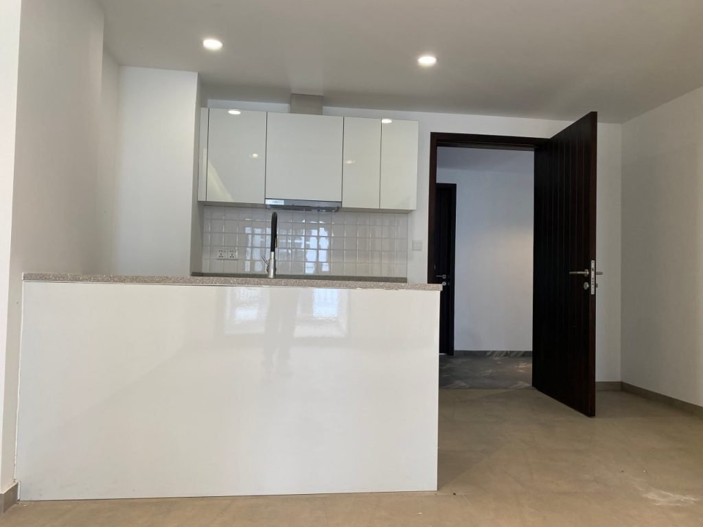 A modern, minimalistic kitchen with white cabinets, a tiled backsplash, and a small countertop. The area is well-lit with recessed ceiling lights, and there is an open dark wooden door on the right.