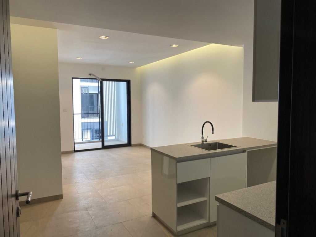 A modern, unfurnished apartment interior with beige floor tiles, a kitchen with a sink and open shelving, and large glass doors leading to a balcony with a city view.