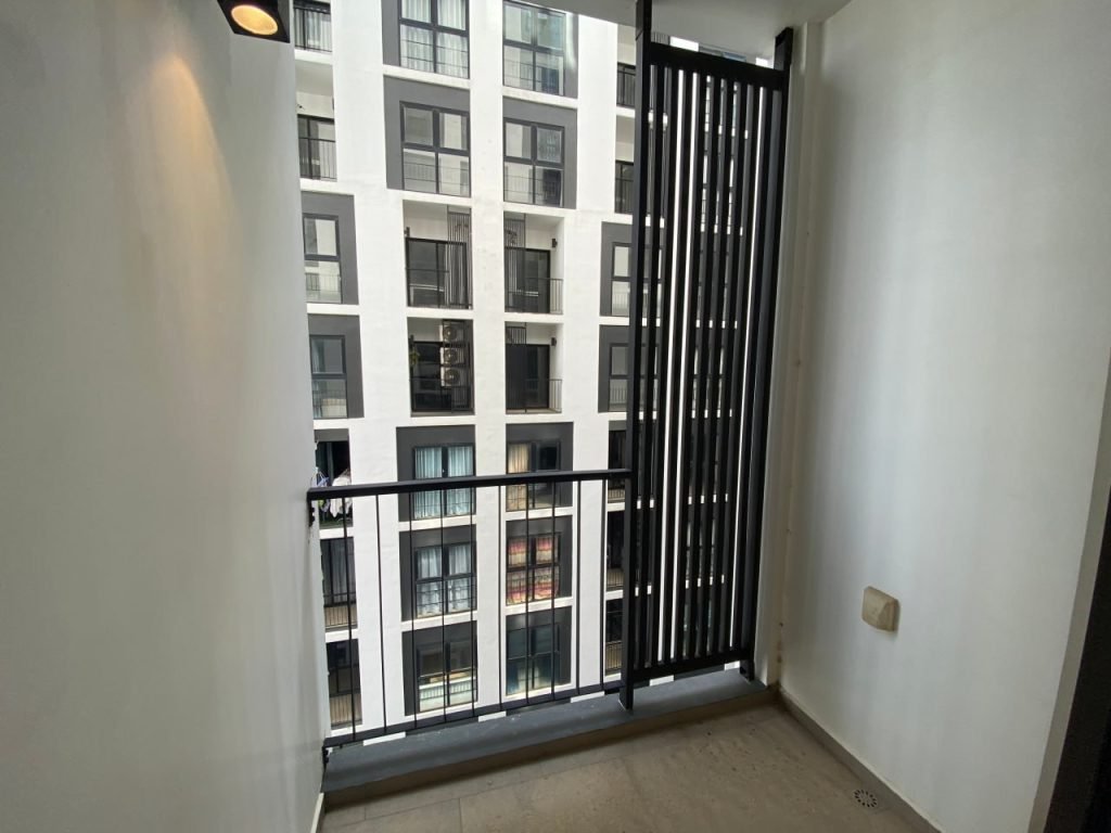 A small, empty balcony with a black railing overlooks a modern apartment building with many windows and balconies. One side of the balcony has vertical black slats; the walls and floor are neutral-colored.