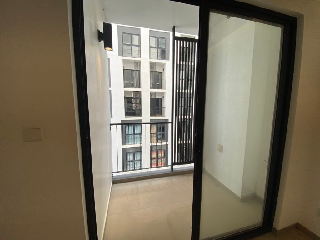 View through a sliding glass door onto a small, modern balcony with a black railing and wall lamp; across the way, the exterior of a neighboring apartment building with windows is visible.
