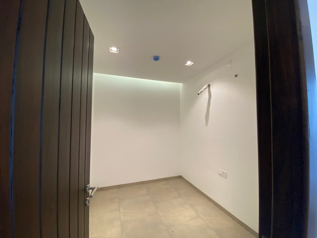 A small, empty room with beige tiled flooring, white walls, and a partially open wooden door. Ceiling lights are on, and a blue smoke detector is visible. There is an exposed pipe or conduit on the right wall.