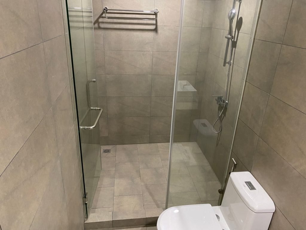 Modern bathroom with beige tiled walls and floor, featuring a glass-enclosed walk-in shower and a white toilet. A metal towel rack is mounted on the wall inside the shower area.