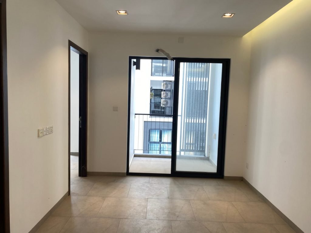A bright, empty room with tiled floors, white walls, a doorway on the left, and large glass doors leading to a small balcony with a metal railing and a view of neighboring buildings.